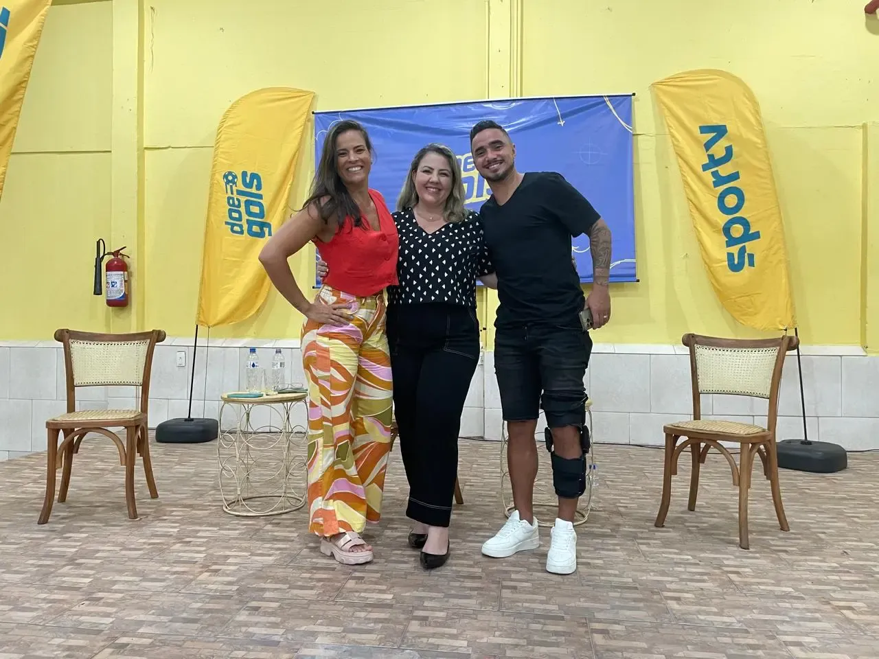 Três participantes posam para foto em frente ao backdrop do Doe Gols e bandeiras do SporTV, durante o evento Doe Gols Educação no clube Ascaer, na Ilha do Governador.