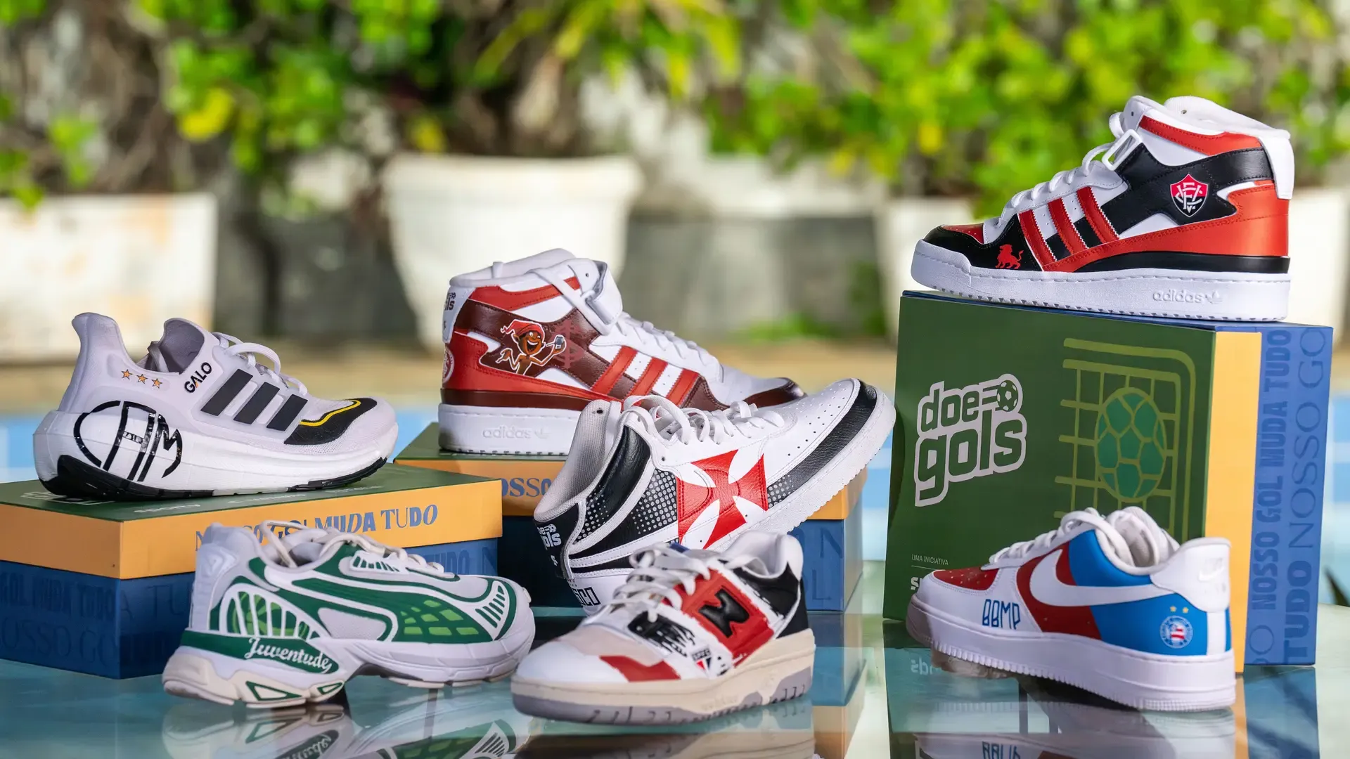 Coleção de tênis personalizados do Doe Gols com as cores e escudos de Atlético-MG, Bahia, Internacional, Juventude, São Paulo e Vitória, dispostos sobre as caixas do projeto com o slogan "Nosso Gol Muda Tudo".