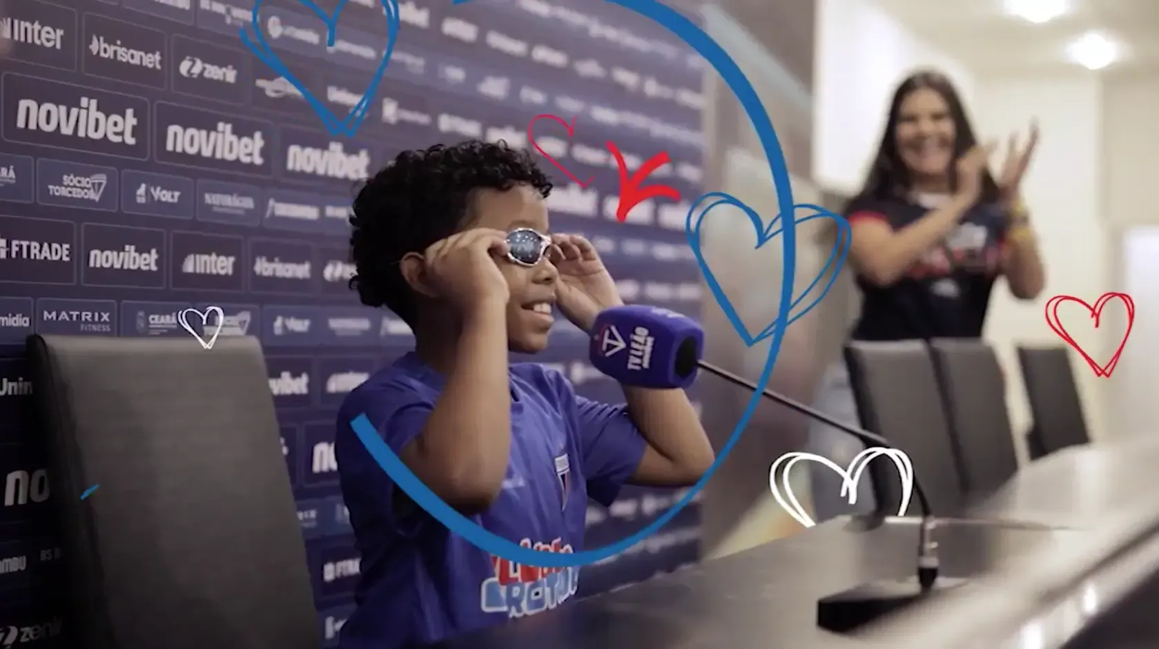 Criança experimenta óculos de sol na sala de coletiva do Castelão, com corações desenhados ao redor, durante a visita ao estádio pelo Jogo Doe Gols do Fortaleza.