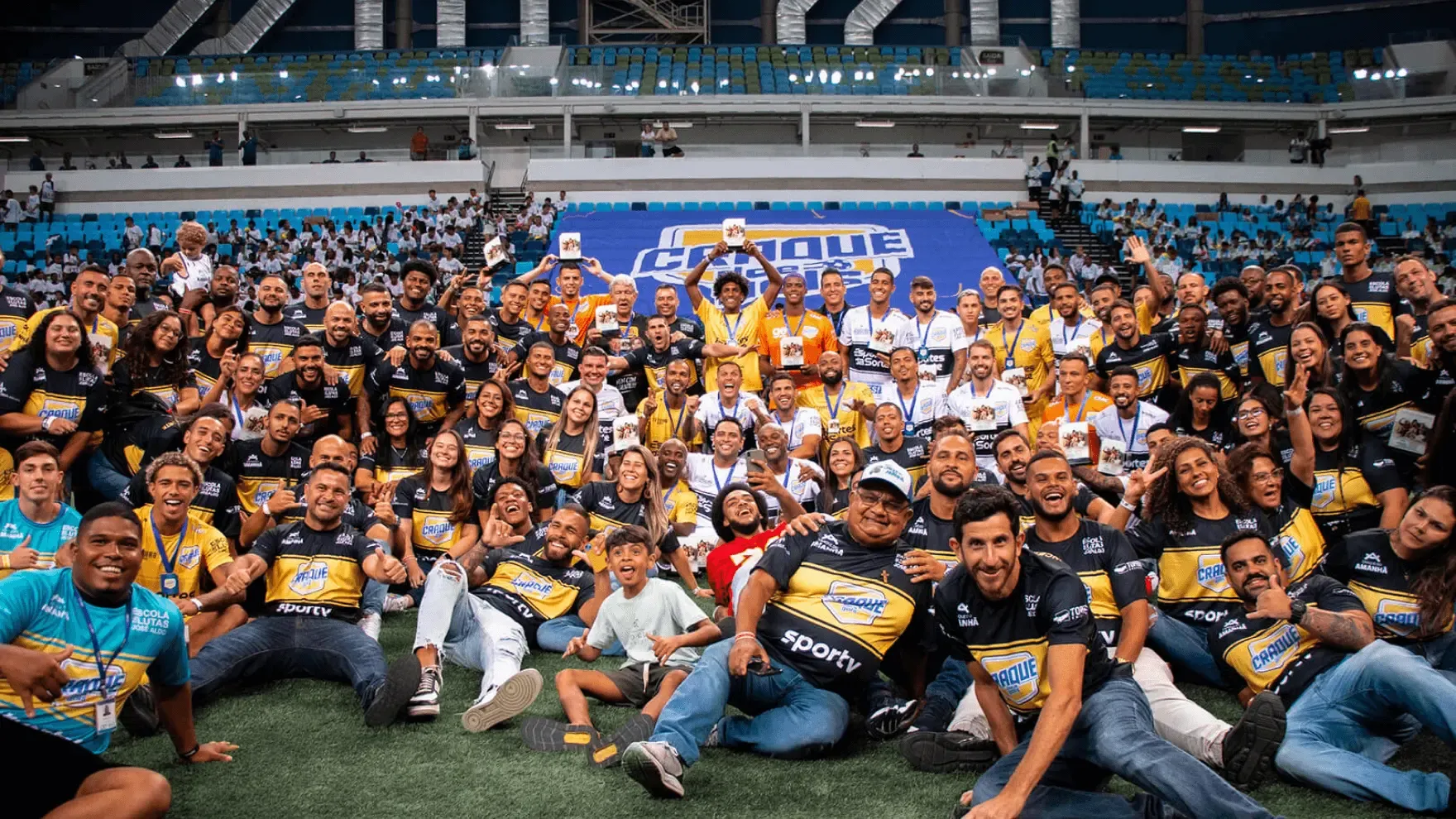 Foto em grupo de participantes do evento "Craque de Amanhã" em um estádio. Dezenas de pessoas posam sorrindo no gramado, usando camisetas pretas e amarelas com os logos "Craque" e "SporTV". Ao fundo, um telão exibe o nome do evento e as arquibancadas azuis do estádio.