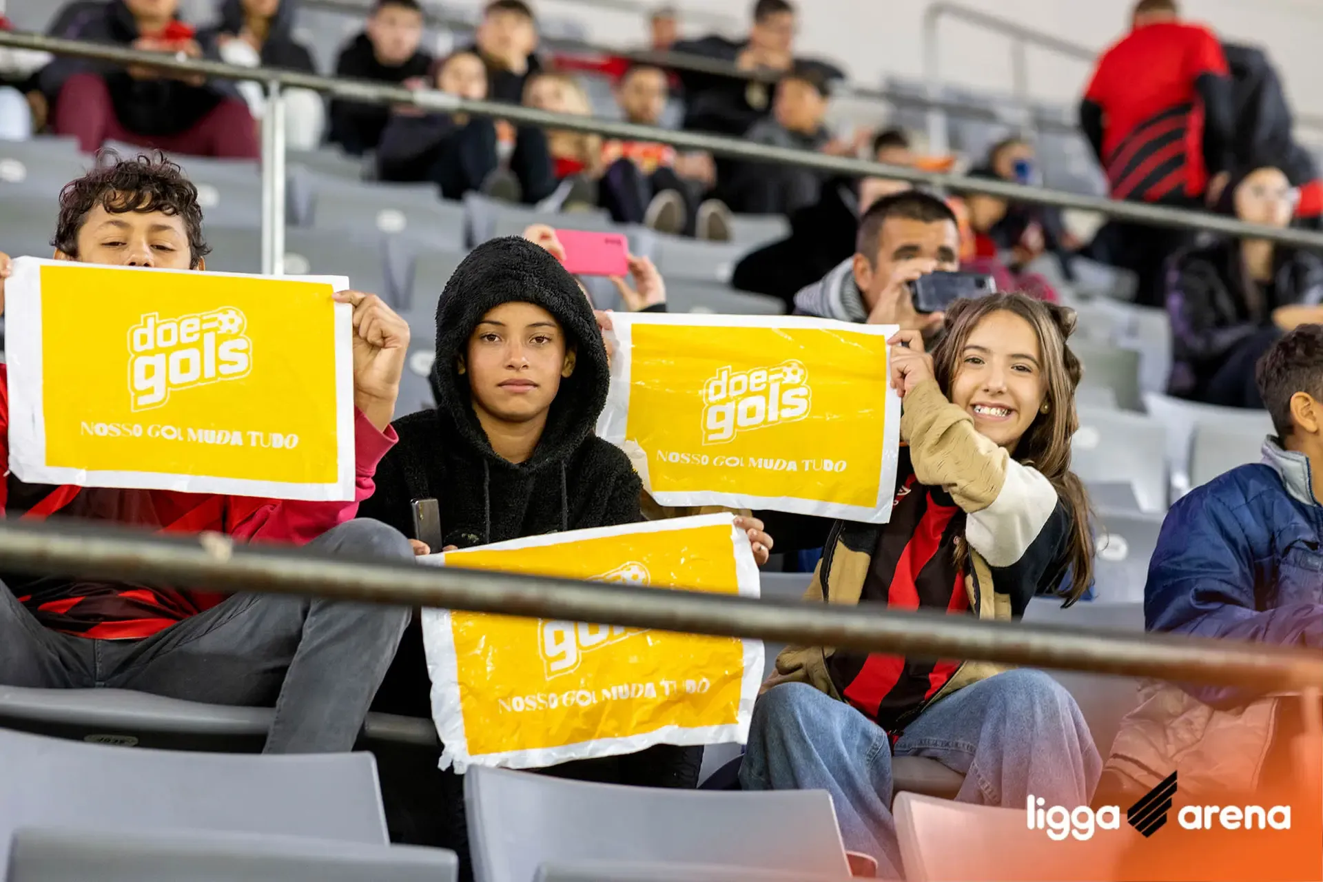 Jovens nas arquibancadas da Ligga Arena exibem bandeirolas amarelas do Doe Gols com o slogan "Nosso Gol Muda Tudo", durante o Jogo Doe Gols entre Athletico-PR e Bahia.