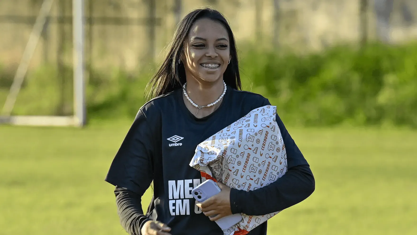 Jovem mulher sorrindo em um campo de futebol, usando camiseta preta da Umbro, colar de pérolas e aparelho dentário. Ela segura um celular e uma sacola da loja Centauro. Ao fundo, uma trave e vegetação desfocada.