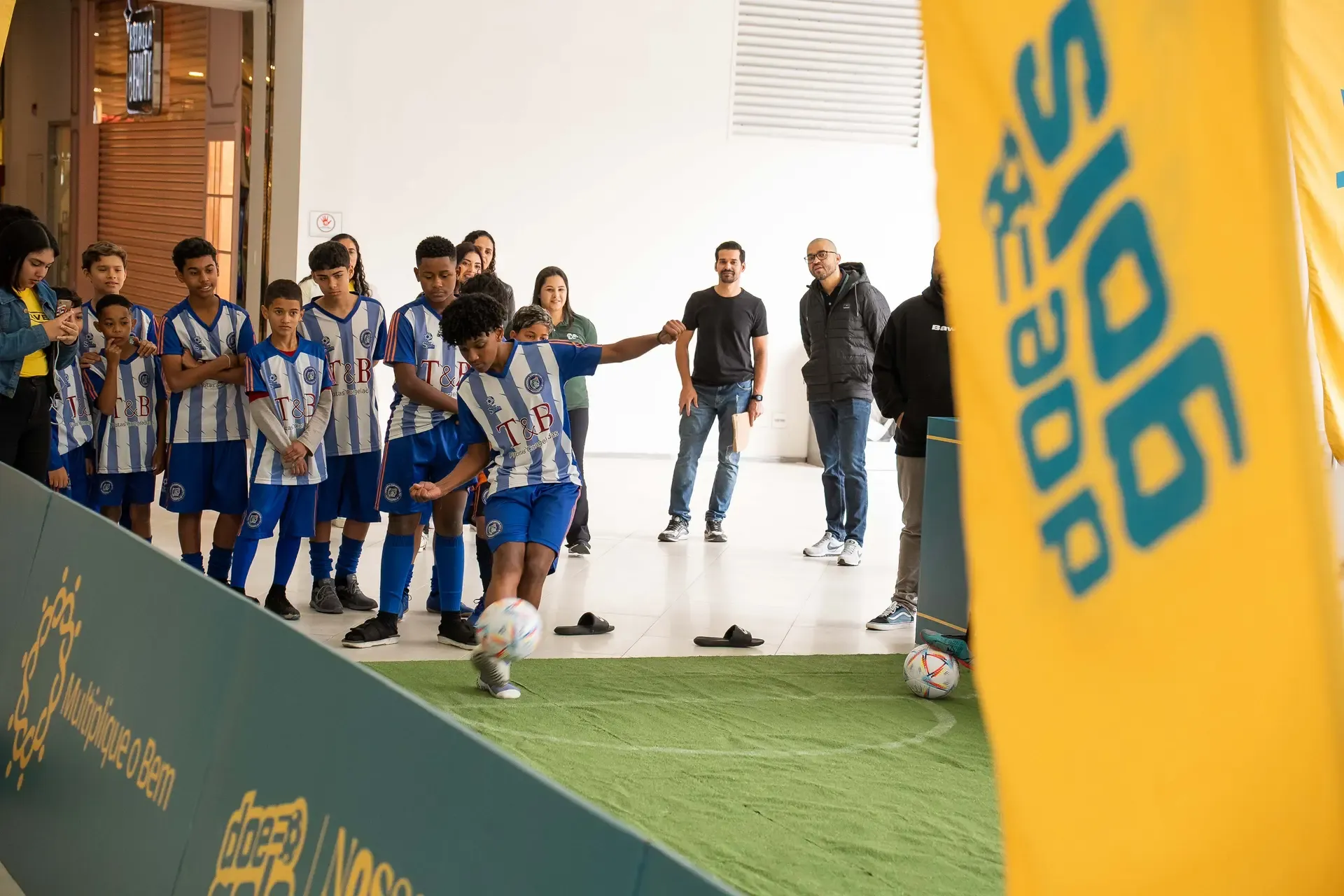 Criança chuta uma bola em gol inflável do Doe Gols no MorumbiShopping, enquanto colegas com uniforme de futebol aguardam sua vez ao fundo.